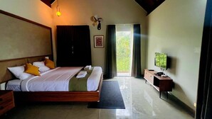 A hotel room with a large bed, a TV, a desk, and a view of greenery outside.