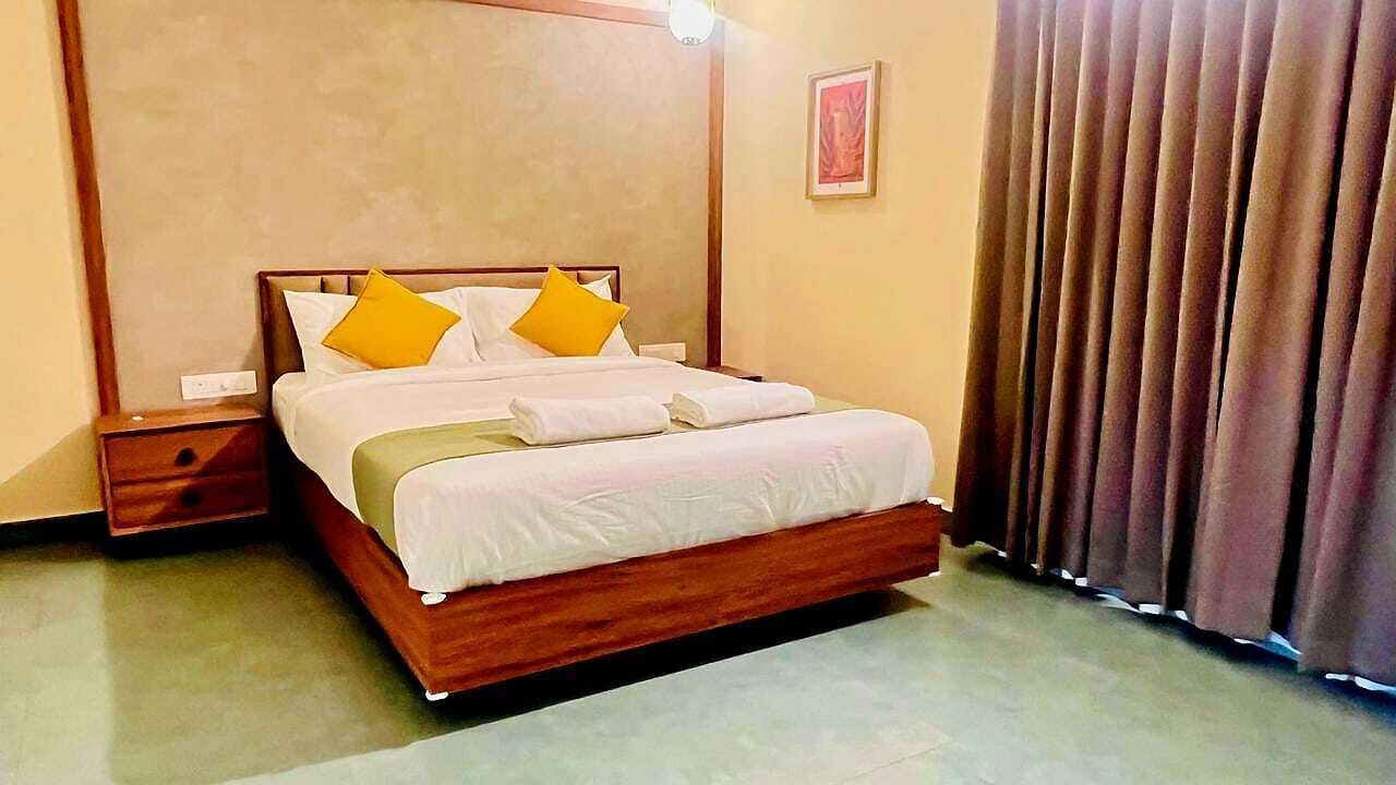 A neatly made bed with white linens and two yellow pillows, a wooden headboard, a bedside table, and a window with curtains.