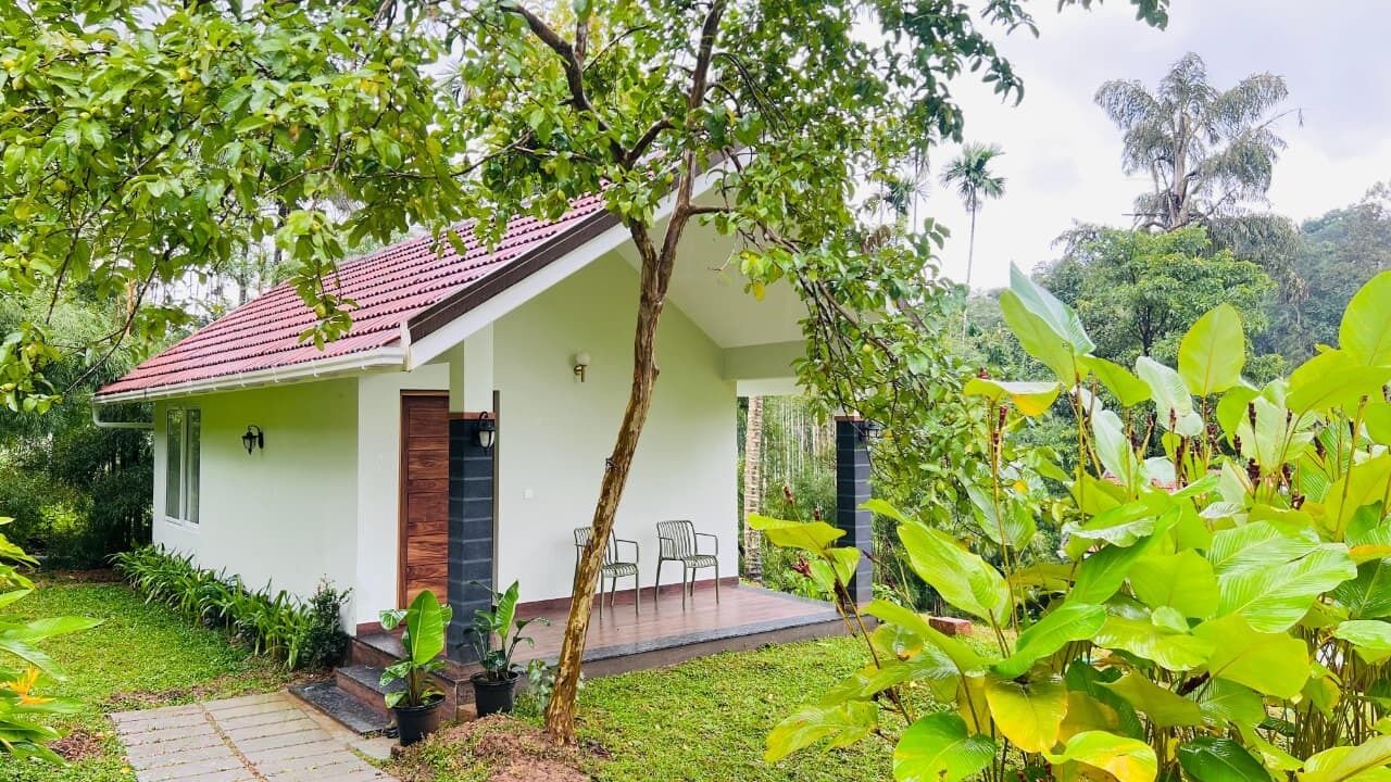 A small, single-story house with a red-tiled roof, a front porch with two chairs, and a garden with various plants.