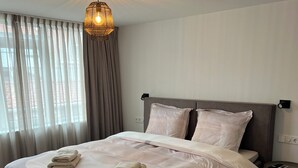En pænt redt seng med gråt sengegavl og hvide sengetøj, et lille natbord med en telefon, og en hængende lampe over sengen.