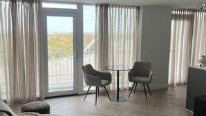 En moderne stue med en grå sofa, to lænestole, et lille rundt bord og et tv-møbel. Store vinduer med lette gardiner giver udsigt til det fri.