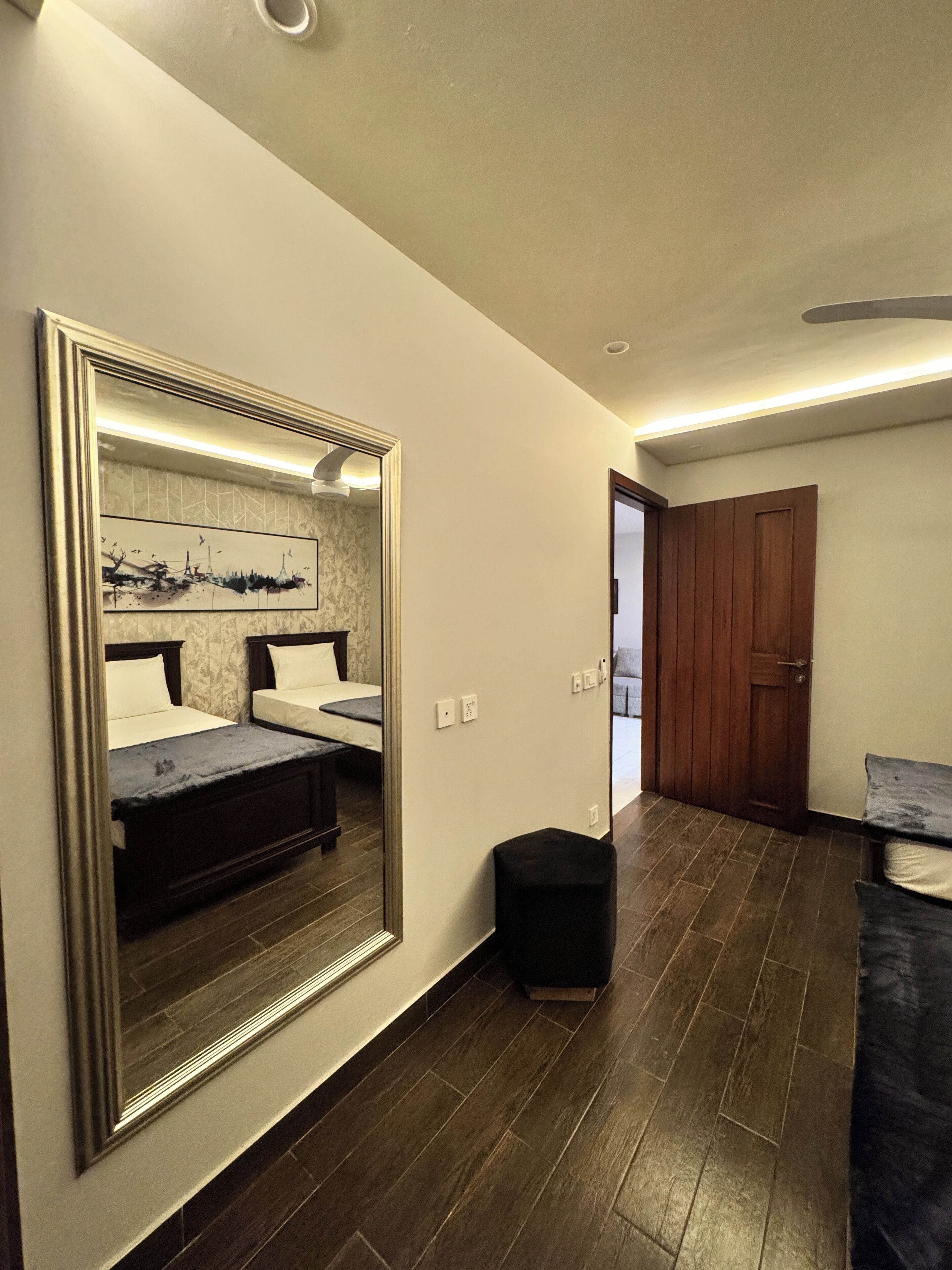 A hotel room with a large mirror, a bed, a small stool, and a wooden door.