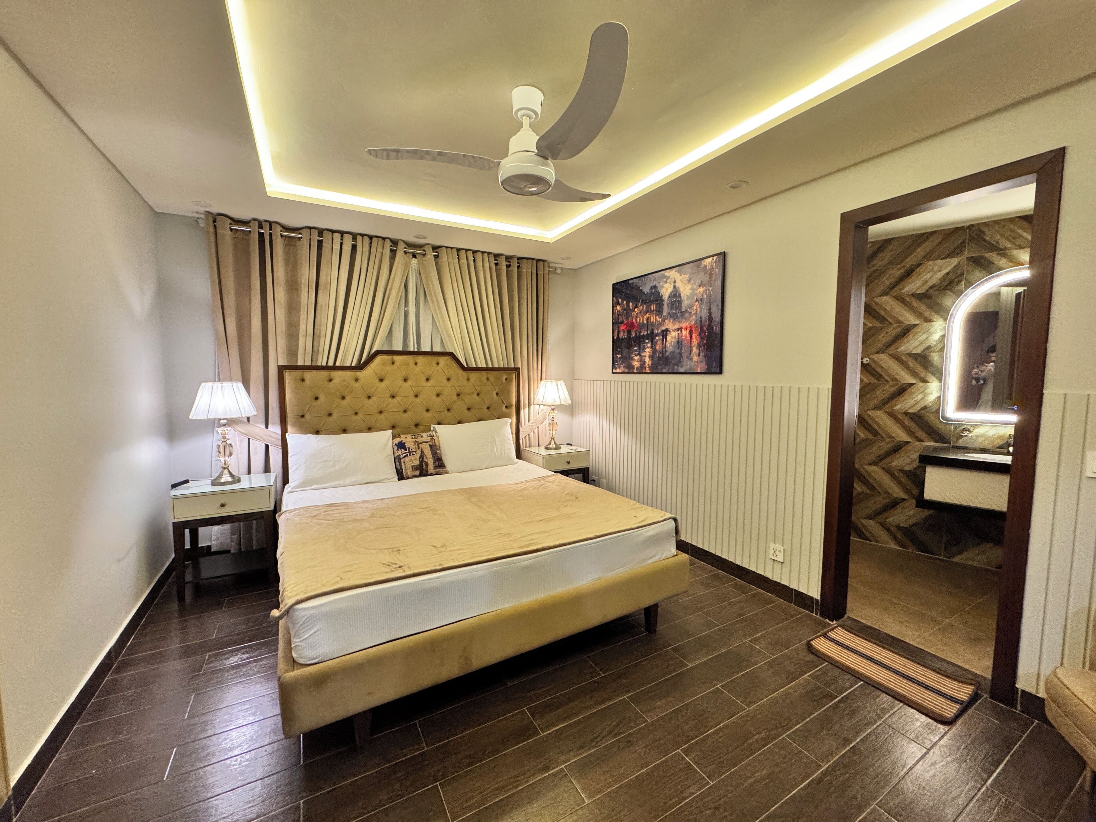 Habitación de hotel con una cama grande, dos mesitas de noche con lámparas, un ventilador de techo, un cuadro en la pared y un espejo en el umbral.