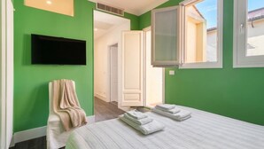 A bedroom with a bed, a TV mounted on the wall, a window, and a door leading to another room.