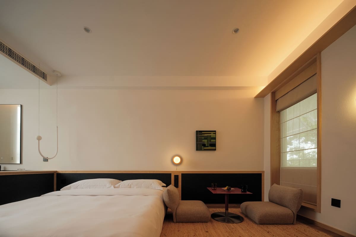 A hotel room with a large bed, a small table, a chair, a wall-mounted lamp, and a window with blinds.