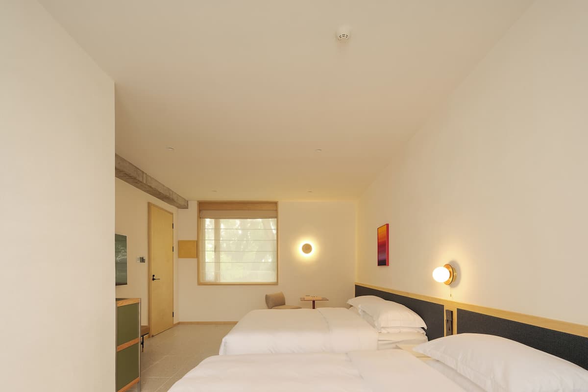 A hotel room with two beds, a small table, a chair, a window with blinds, and a wall-mounted light fixture.