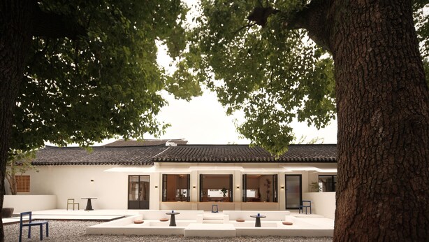 Un edificio de estilo tradicional con tejado de baldosas, un patio con asientos y un gran árbol en primer plano.