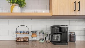 A kitchen with a Keurig coffee maker, a coffee grinder, a jar of coffee, and a plant on the counter.