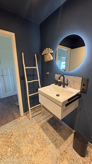 A bathroom with a white sink, dark blue walls, a wooden ladder, a round mirror, and a decorative basket.