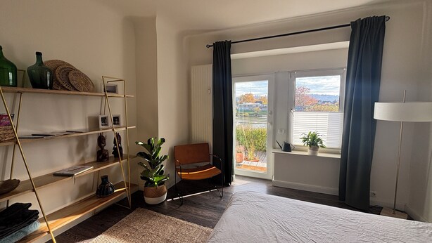 A bedroom with a bed, a chair, a window with curtains, a shelf with decorative items, and a view of the outdoors.