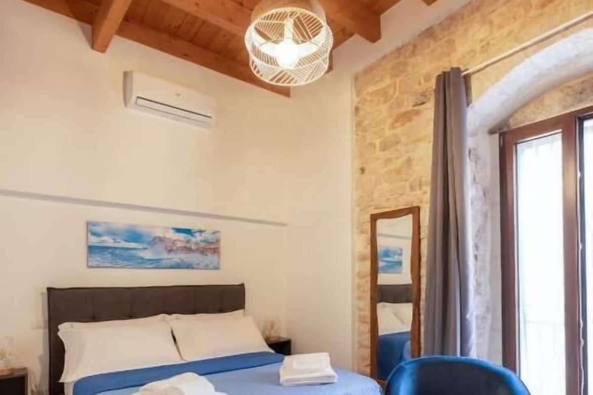 A bedroom with a bed, a blue chair, a hanging light fixture, a painting, and a stone wall.