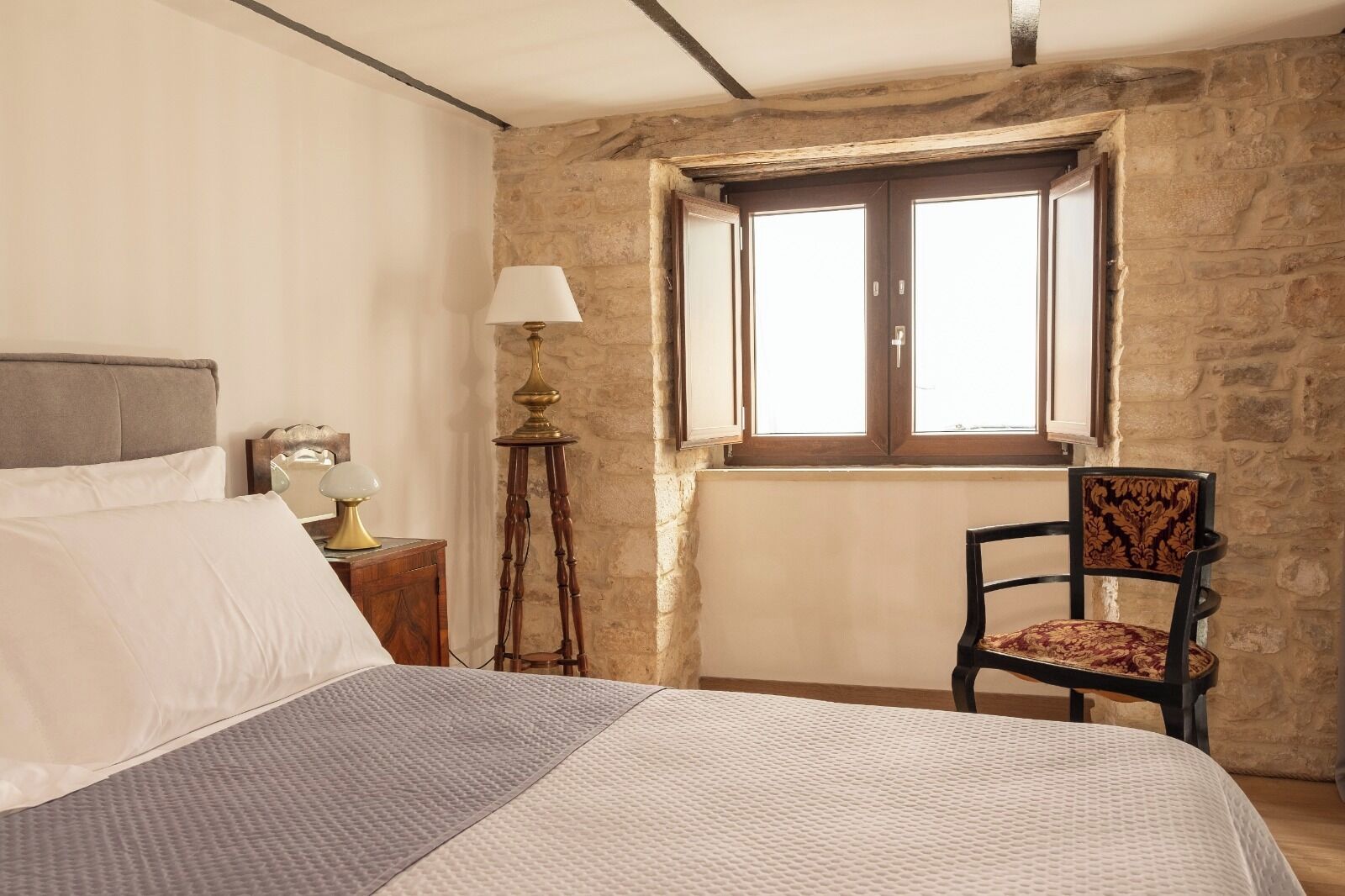 A bedroom with a stone wall, a bed with a grey and white duvet, a wooden nightstand with a lamp, and a chair with a patterned cushion.