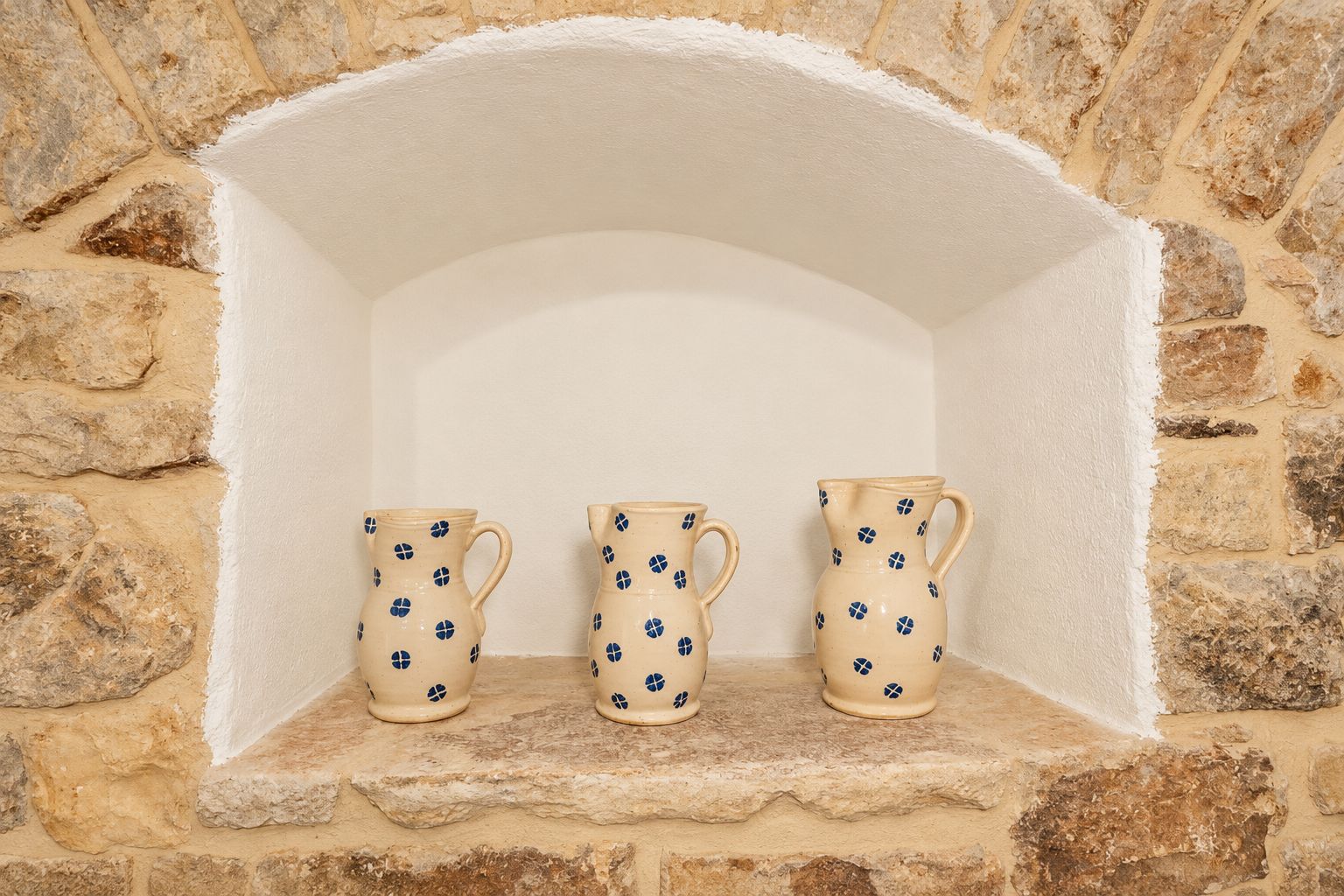 Three ceramic pitchers with a blue pattern, placed on a stone shelf against a stone wall.