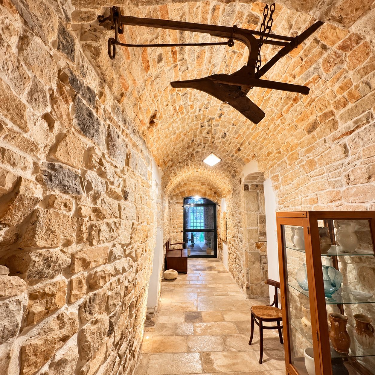 A stone-walled corridor with a hanging light fixture, a glass display case with pottery, and a chair.