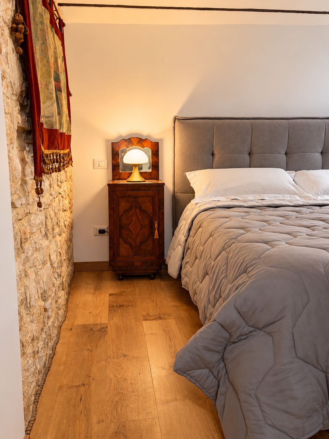 A bedroom with a bed, a nightstand with a lamp, a stone wall, and a wooden floor.
