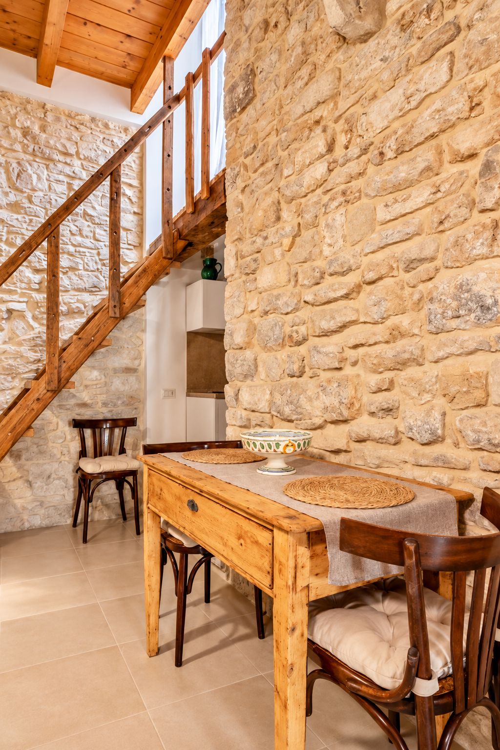 A rustic interior with a stone wall, wooden staircase, and a dining table with chairs.