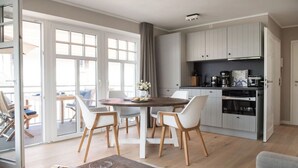 A modern kitchen with white cabinets, a dining area with a round table and chairs, and a view through large glass doors to an outdoor seating area. - Comfortable vacation apartment Meertied in the Lindenhof on Wangerooge with balcony (Wangerooge)