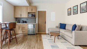 A modern kitchen with wooden cabinets, a microwave, and a coffee machine. There is a dining area with two chairs and a table. A sofa and a coffee table are in the living area. - Aurora Lite - Brand new suite with private entrance (Canmore)