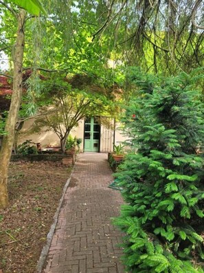 A paved pathway leading to a building with a green door, surrounded by lush greenery and trees. - B&B ARCADIA (Casanova Lonati)