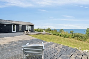 A wooden deck with a bench, a house with a dark exterior, and a view of the water. - “Magical Sea View – From Terrace to Horizon” (Svaneke)