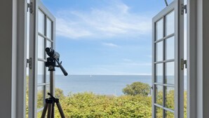 Een open deur met een telescoop op een statief, met uitzicht op een kustlandschap met een houten terras en begroeiing.