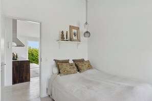 A bedroom with a bed, pillows, a shelf with decorative items, a hanging light, and a view of the outdoors.
