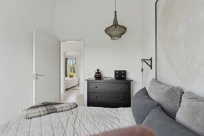 A bedroom with a bed, a dresser, a hanging light fixture, and a view of another room.