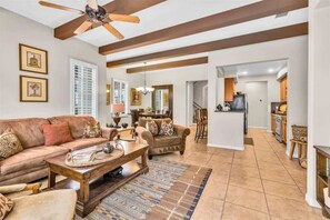 A spacious living room with a large sofa, coffee table, and a ceiling fan. There is a kitchen area visible in the background with wooden cabinets and a dining table. - Desert Paradise | 3BR Legacy Villa (La Quinta)