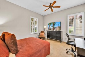 A bedroom with a bed, a desk, a chair, a ceiling fan, a TV, and two windows with blinds.