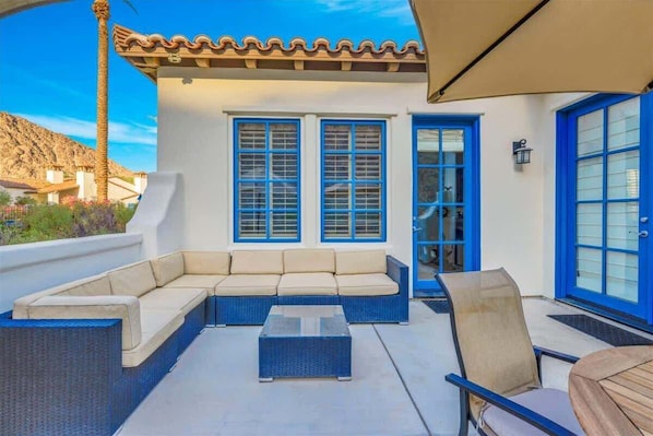 An outdoor patio area with a beige sofa, a glass-top coffee table, and a chair. There are two blue-framed windows and a door with a glass panel.