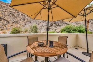 Een terras buiten met een ronde houten tafel, beige stoelen en een grote beige parasol die schaduw biedt.