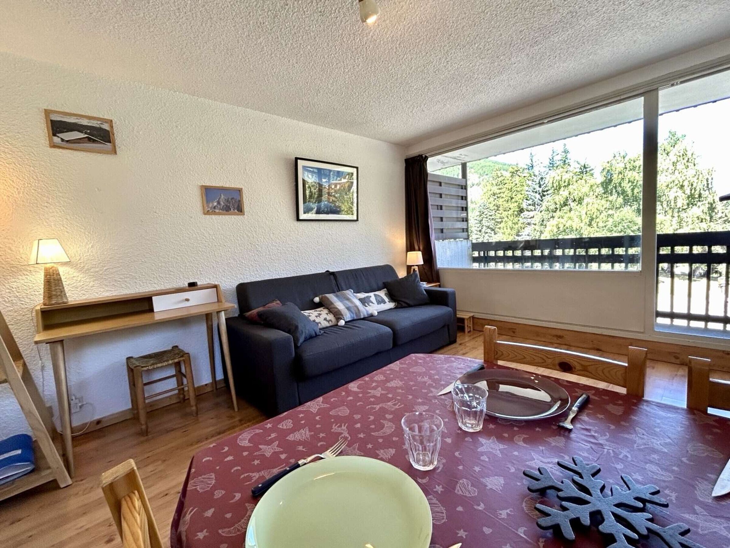 Un salon chaleureux avec un canapé, une table à manger recouverte d’une nappe rouge et une fenêtre donnant sur des arbres.