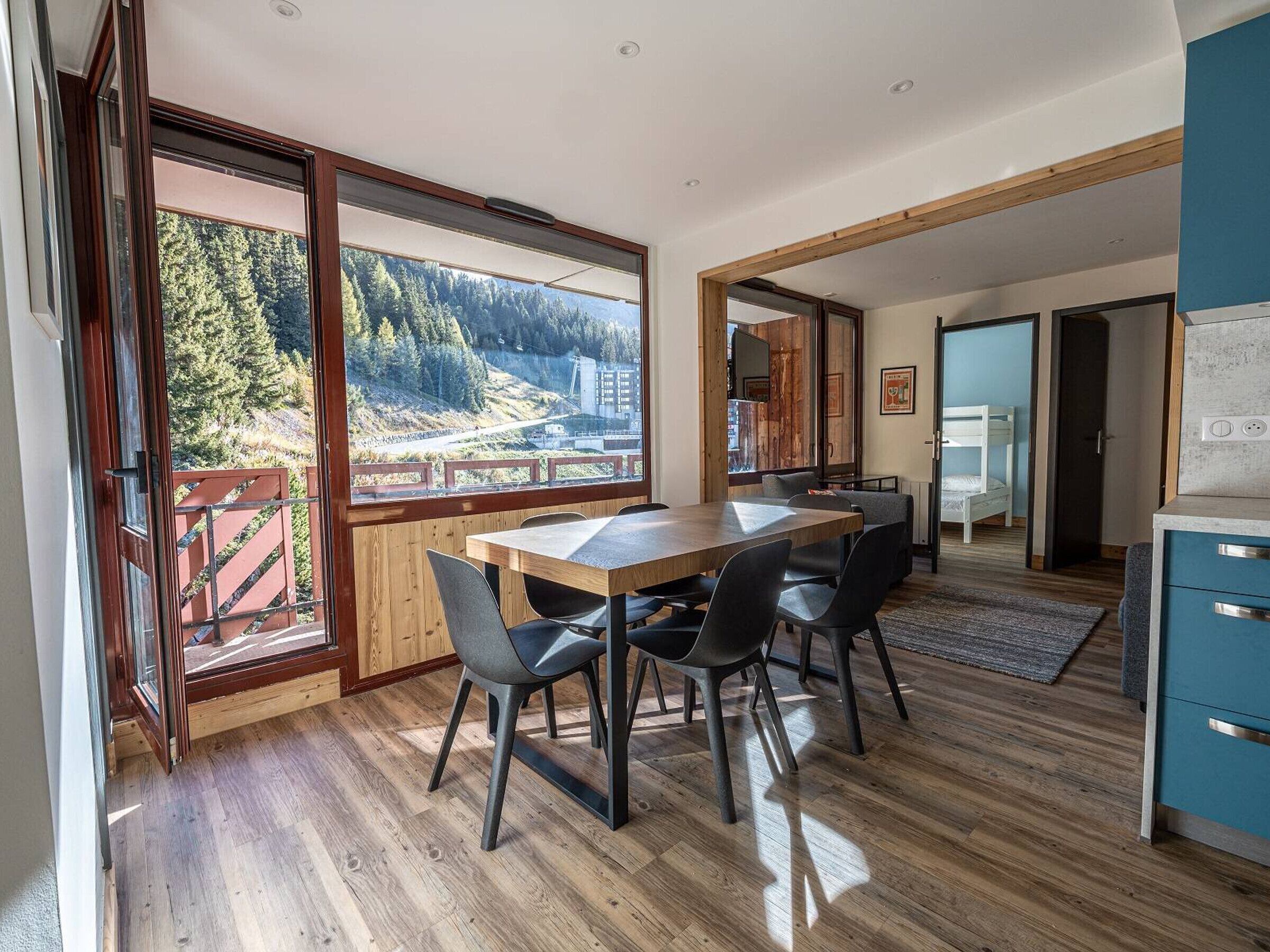 A modern kitchen with wooden flooring, a dining area with a table and chairs, and a large window offering a view of a forested area.