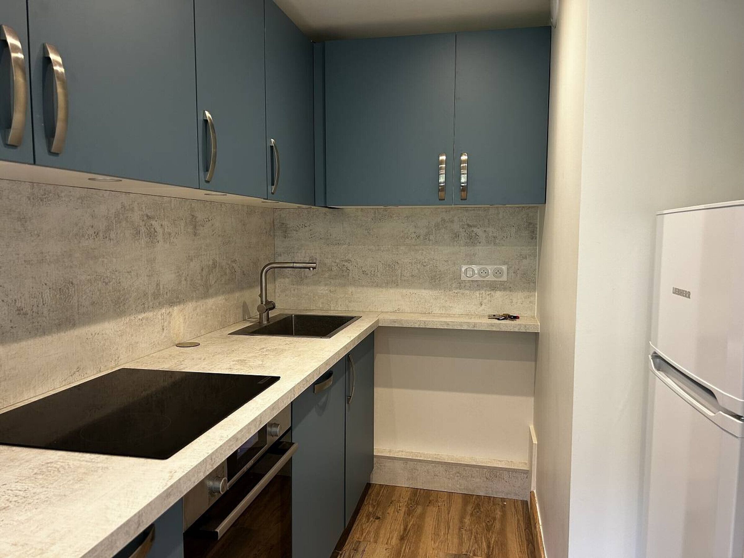 Eine kleine Küche mit blauen Schränken, einer schwarzen Spüle und einem weißen Kühlschrank.