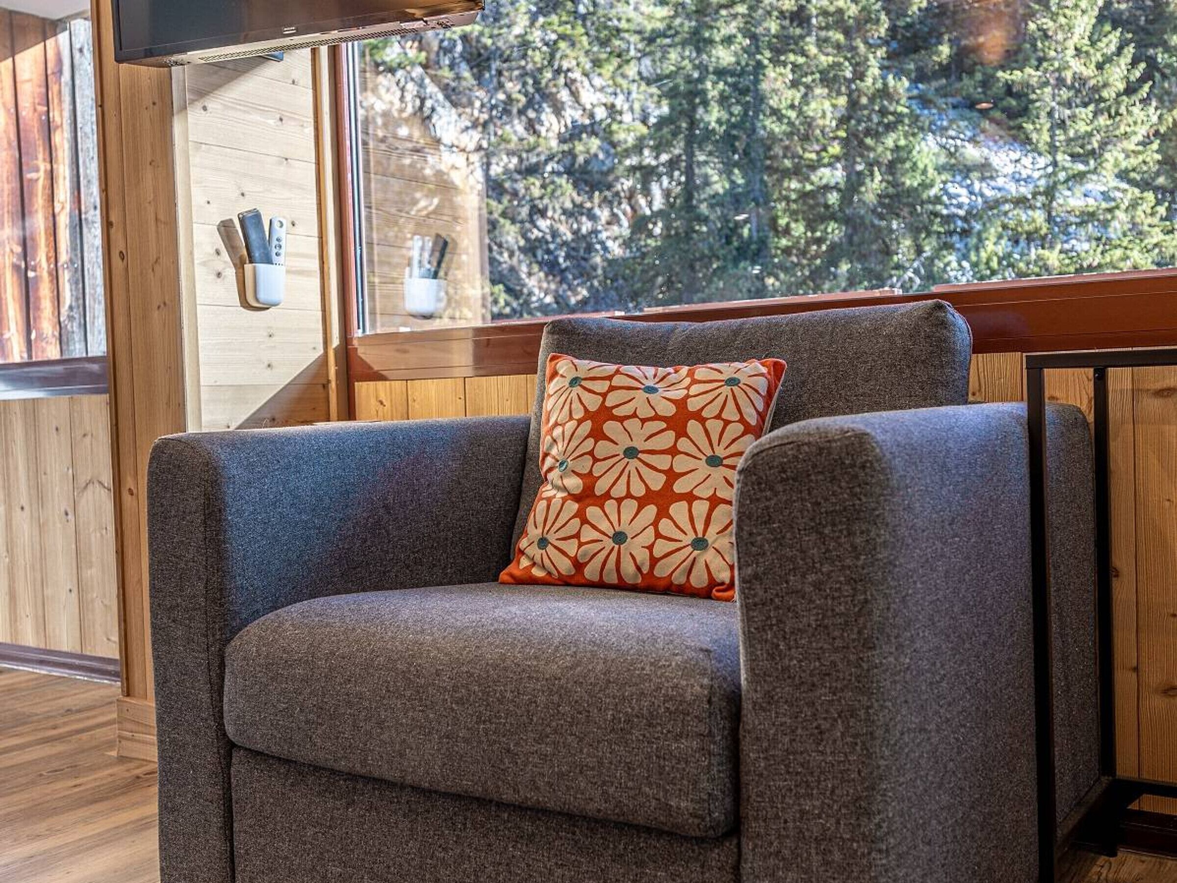 A gray armchair with a floral pillow, a wooden wall, and a window with a view of trees.