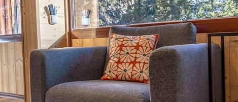 A gray armchair with a floral pillow, a wooden wall, and a window with a view of trees.