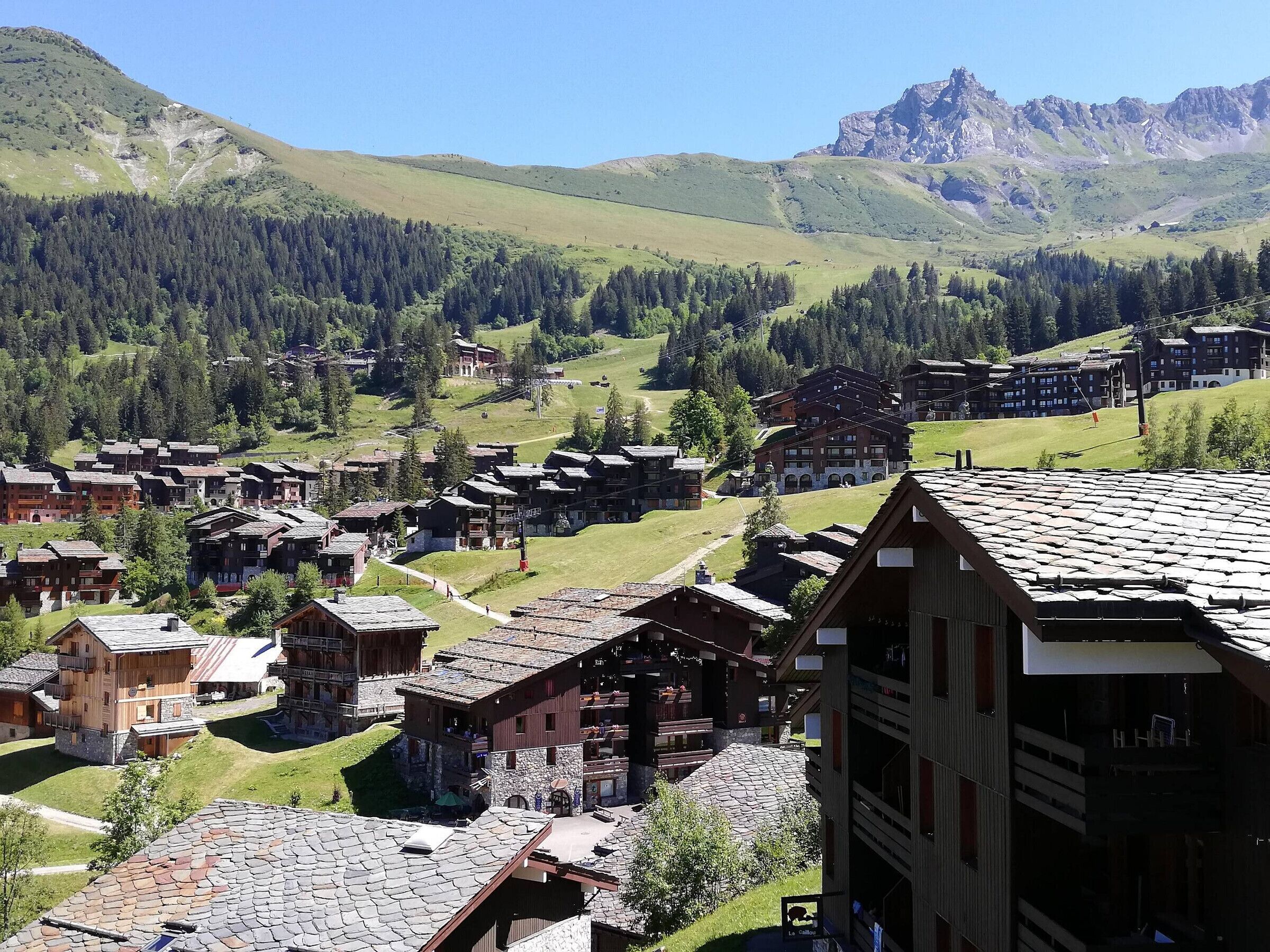 A mountainous village with traditional wooden buildings, a clear sky, and a view of distant rocky mountains.