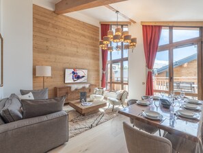 Un salon moderne avec un plafond en bois, un coin repas doté d’une table en verre et de grandes fenêtres ornées de rideaux rouges.