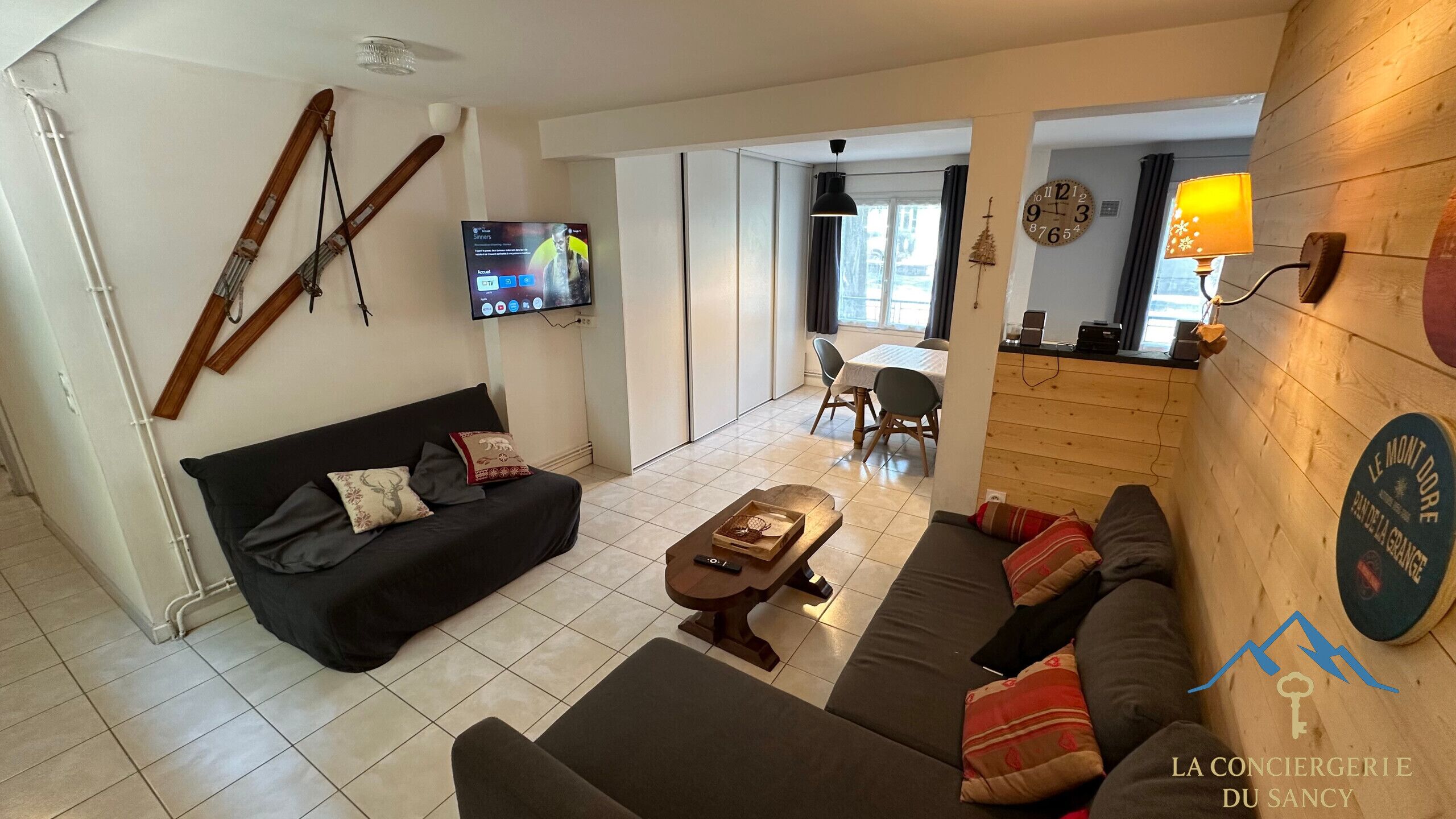 Un salon moderne avec un canapé noir, une table basse en bois et un téléviseur fixé au mur. On aperçoit à l’arrière-plan un coin repas avec une table et des chaises.