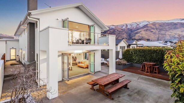 Une maison moderne à deux étages, dotée d’un balcon, d’une terrasse avec une table et des bancs en bois, et offrant une vue sur la montagne.