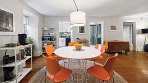 Une cuisine moderne avec des meubles blancs, un coin repas doté d’une table ronde et de chaises orange, et un grand luminaire suspendu.
