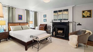 Une chambre à coucher comprenant un lit, des tables de chevet, une chaise, une cheminée et des œuvres d’art accrochées aux murs.