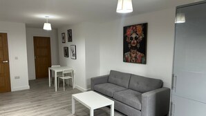 Una sala moderna con un sofá gris, una mesa de centro blanca y una mesa de comedor con sillas. Hay cuadros enmarcados en la pared y un cuadro grande sobre el sofá.