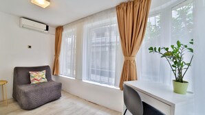 A modern room with a grey sofa, a small table, a desk with a chair, and a potted plant. - Garni House 46 Plus (Belgrade)