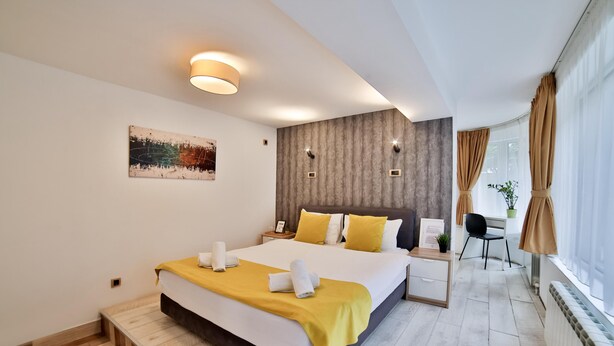 A modern hotel room with a large bed, a desk, and a chair. There is a window with curtains, a plant, and a painting on the wall. - Garni House 46 Plus (Belgrade)