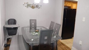 A dining area with a glass table, four chairs, a baby high chair, a black refrigerator, and a decorative wall piece.