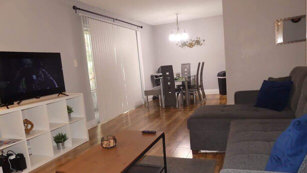 A modern living room with a flat-screen TV, a sofa, a coffee table, and a dining area with chairs and a table.