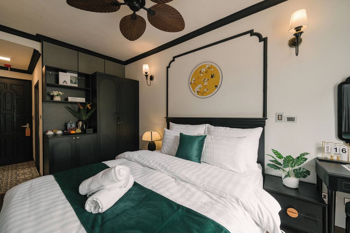 Una cama bien hecha con ropa de cama blanca y verde, una mesita de noche con una planta y un reloj de pared.