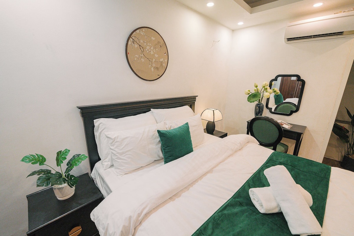 Habitación de hotel con una cama bien hecha, una mesita de noche con lámpara, una planta en maceta y un espejo en la pared.