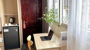 A compact kitchen with a refrigerator, a wooden cabinet, a small table with a laptop, a potted plant, and a window with curtains.